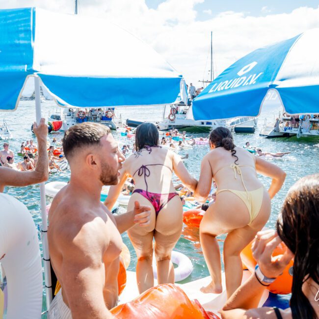 A group of people in swimsuits enjoy a lively boat party, with inflatable floats, umbrellas, and boats on the water under a sunny sky. The scene is vibrant and energetic.