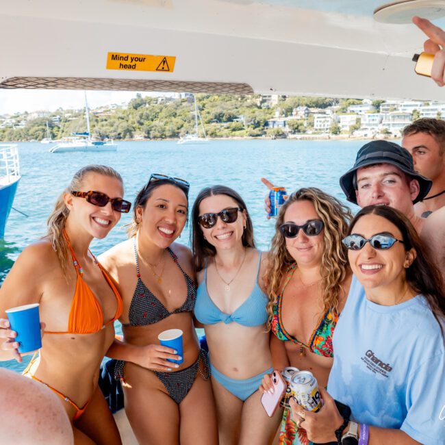 A group of six smiling young adults in swimwear pose together on a boat, holding drinks. The sea and shoreline are visible in the background. A "Mind your head" sign is overhead.