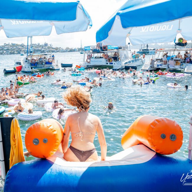 A woman in a bikini sits on a large inflatable at the edge of a crowded lake, surrounded by people on colorful floats and boats under sunny skies. Two blue-and-white umbrellas are overhead.