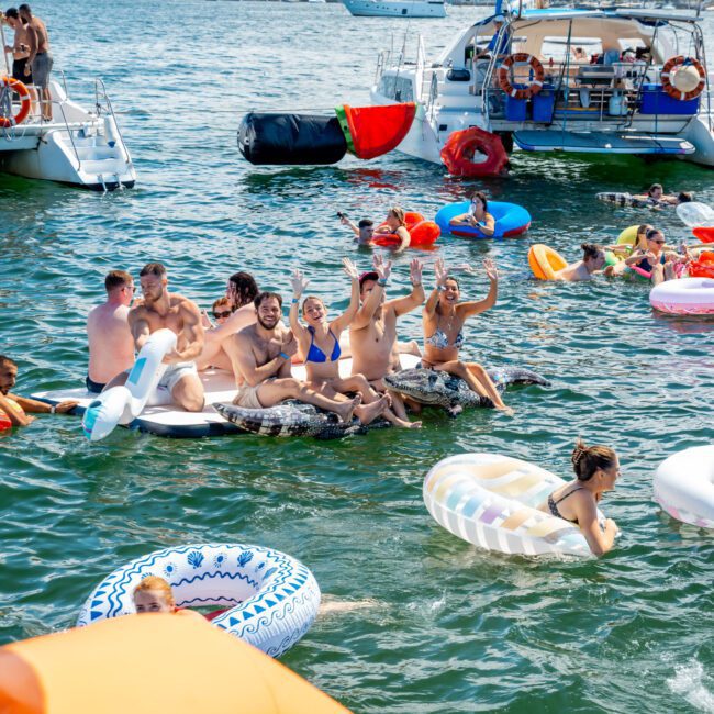 A group of people relax and have fun on inflatable floats in the water near anchored boats on a sunny day. Some wave and smile, surrounded by colorful floaties. The background shows more boats and buildings.