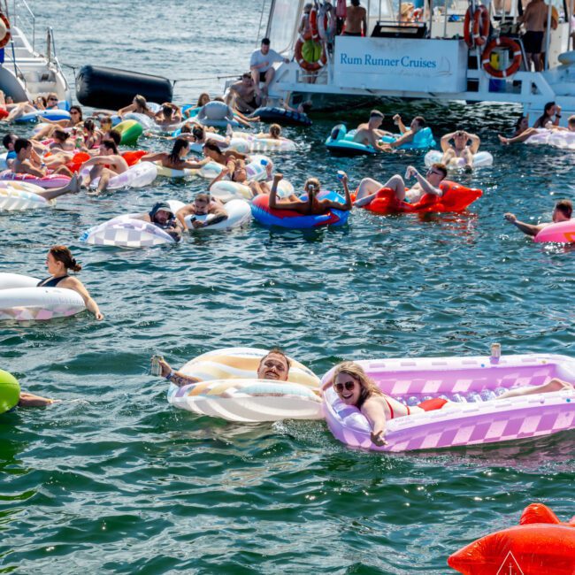 A lively scene of people relaxing on colorful inflatable floats in the water near boats on a sunny day, with a bridge visible in the background and the logo "Yacht Social Club" in the corner.