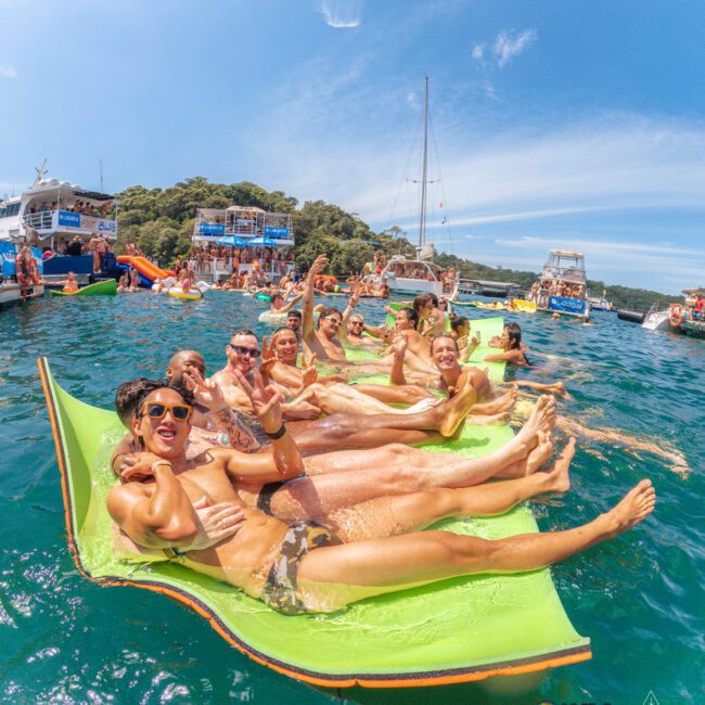 A group of smiling people in swimsuits relax on a floating mat in clear blue water at a lively boat party, with other boats and partygoers in the background under a sunny sky.