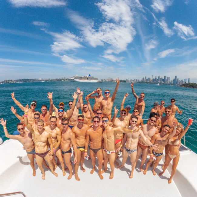 A group of men in swimwear cheerfully pose on a boat under a blue sky, with city skyscrapers and a cruise ship visible in the background. The water is a bright turquoise, and everyone looks happy.