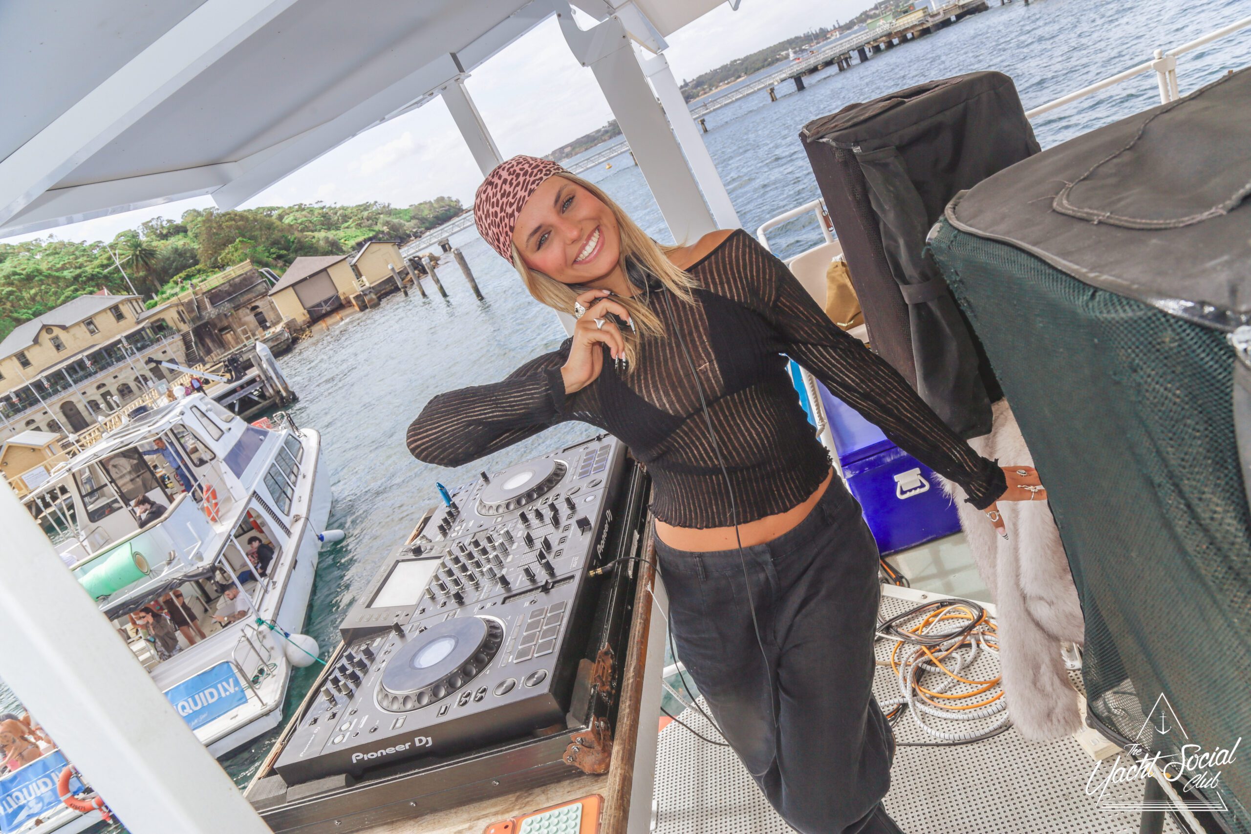 A smiling woman in a black, sheer top and bandana stands next to DJ equipment on a boat, with water, docked boats, and greenery visible in the background.