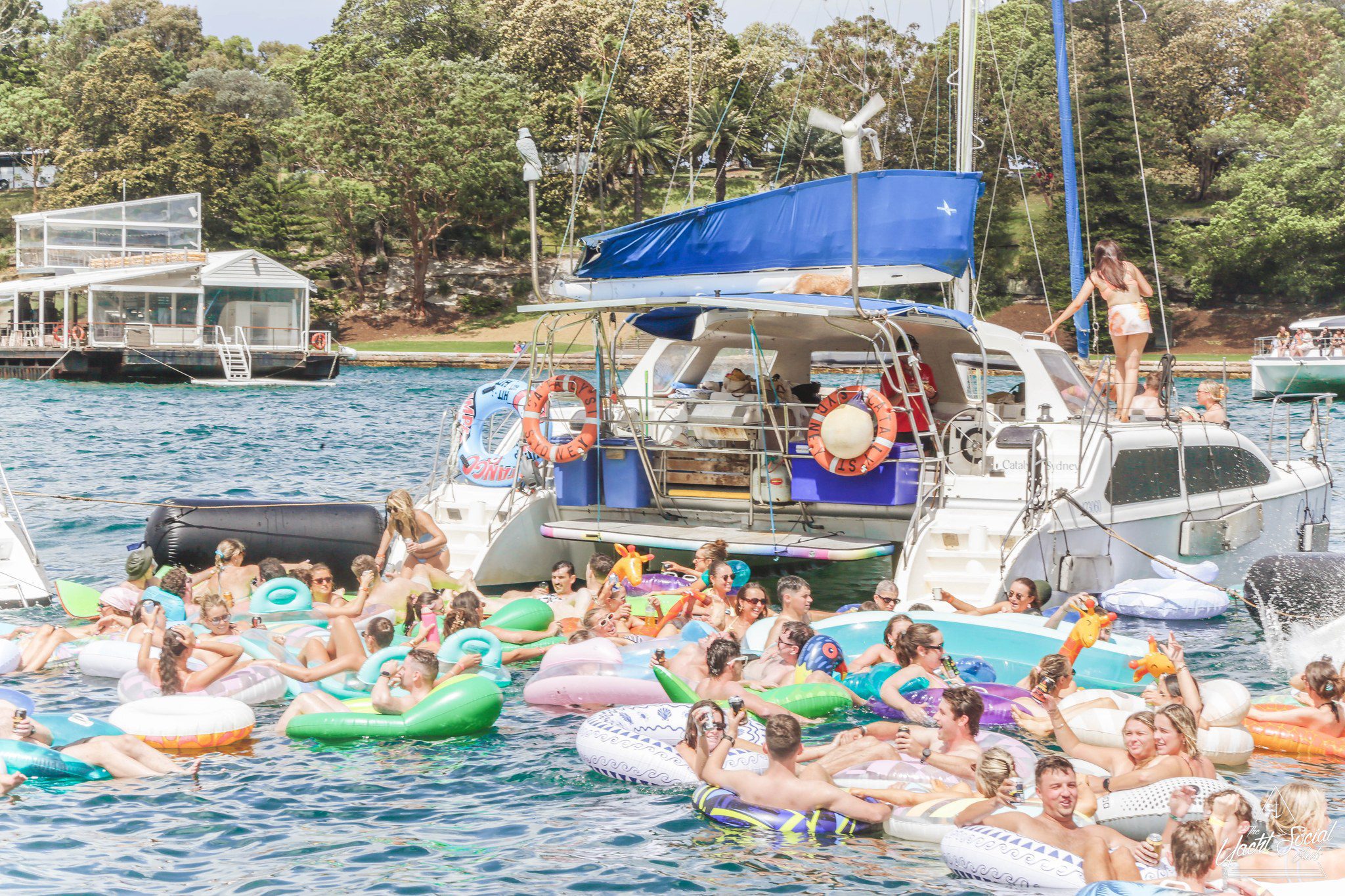 A large group of people relax and socialize on colorful inflatable floats in the water near a yacht, with others on the boat. Trees and more boats are visible in the background on a sunny day.