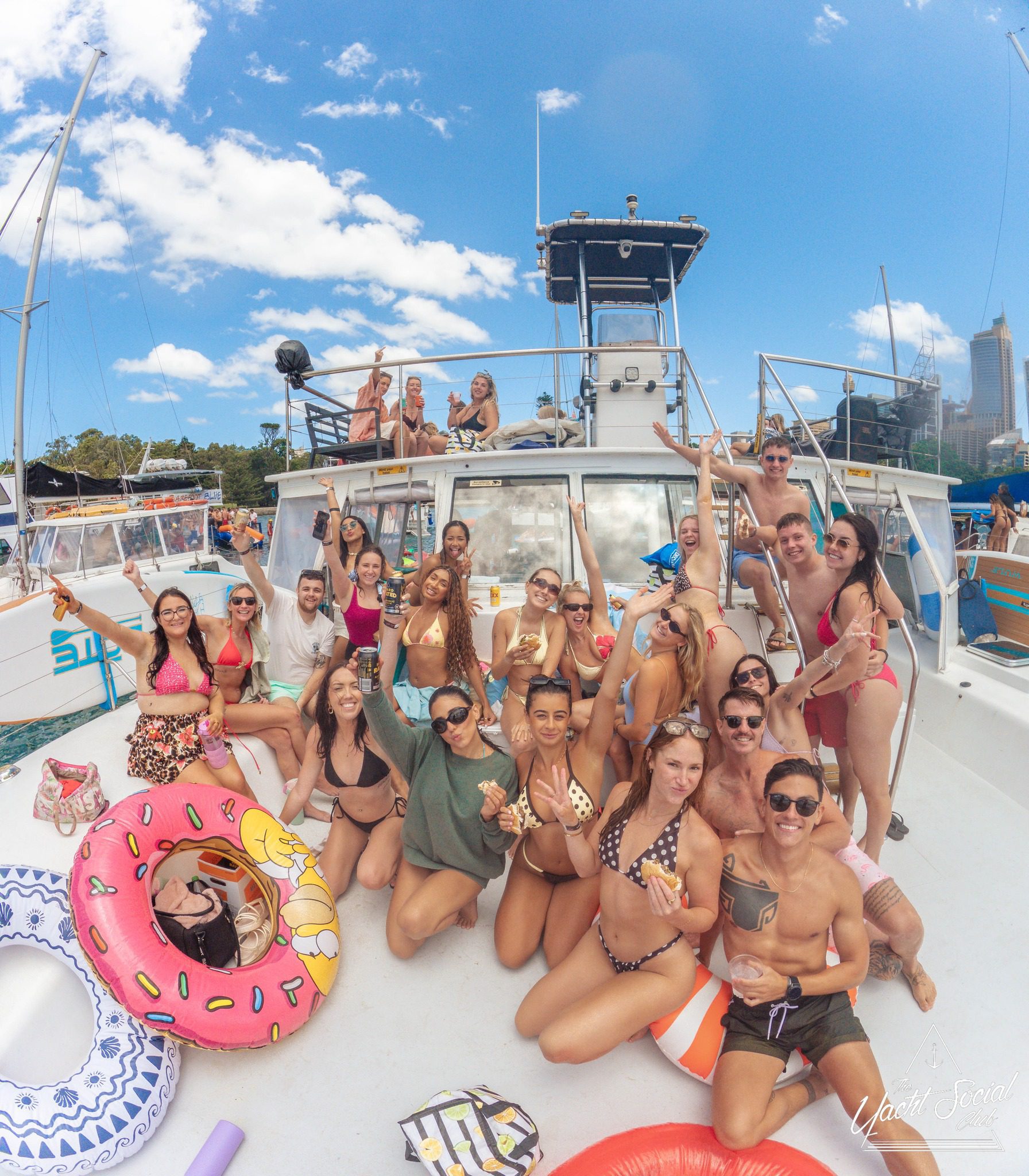 A large group of young adults in swimsuits pose and smile on a boat deck with pool floats, drinks, and sunglasses under a bright blue sky, enjoying a lively party atmosphere.