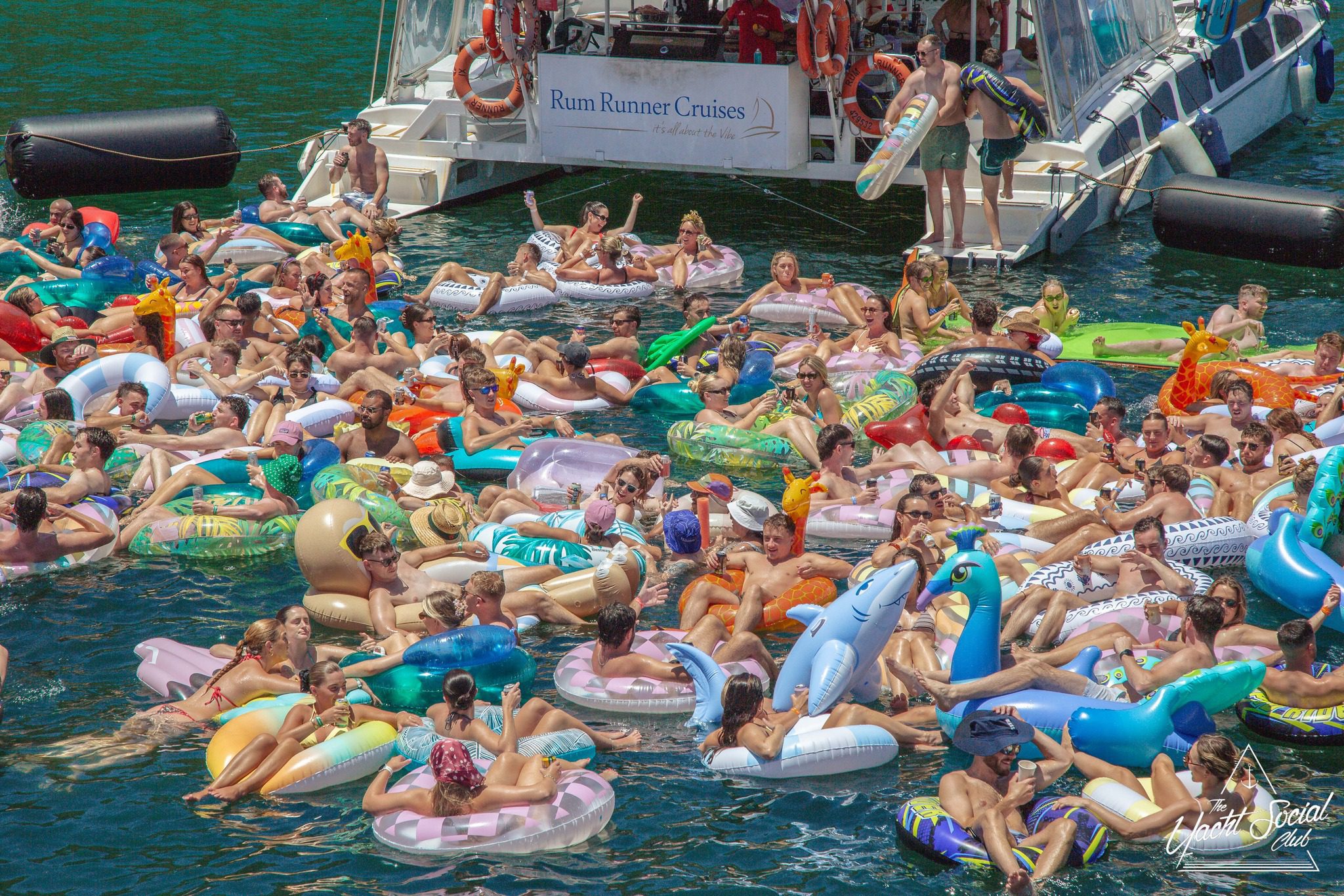 A large group of people in colorful inflatable pool floats gather in the water near a boat labeled "Rum Runner Cruises" on a sunny day, enjoying a lively social event.