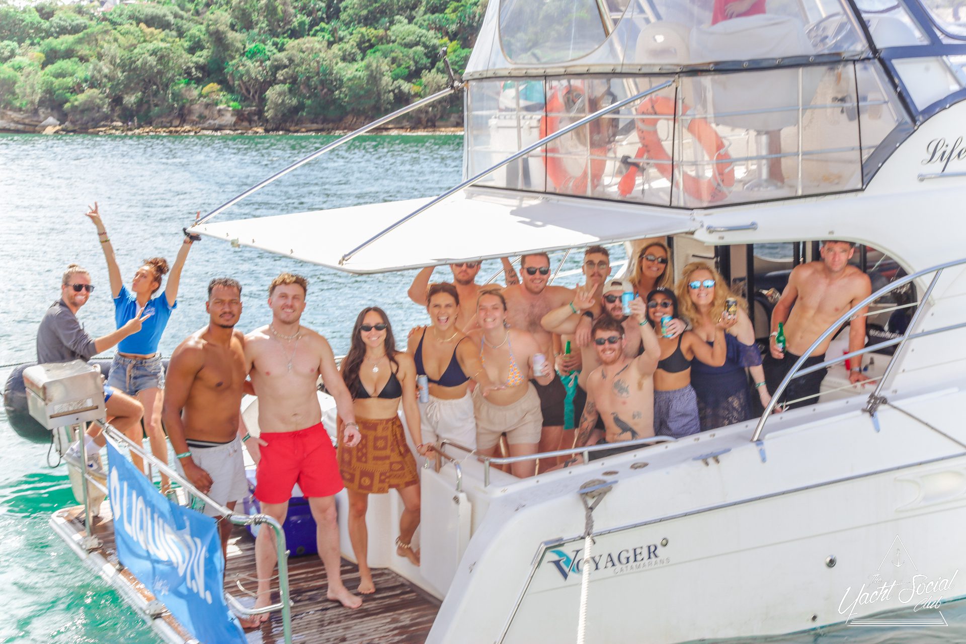 A group of people in swimsuits and summer clothes smile and pose on a white yacht anchored near a lush, green shoreline on a sunny day. Some hold drinks, while others wave or make peace signs.
