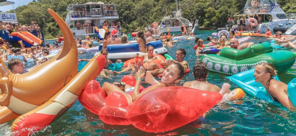 A crowded pool party on a sunny day, with people relaxing on colorful inflatable floats in the water, surrounded by boats and lush green trees—perfect for a Yacht Social or other exciting Yacht events.