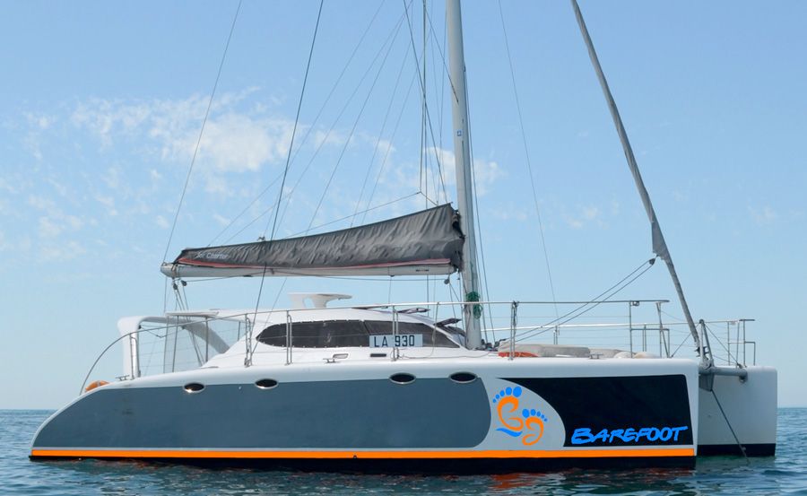 A modern catamaran sailboat with a gray hull and white deck, featuring the name "Barefoot" and an orange foot logo, floating on calm blue water under a partly cloudy sky.